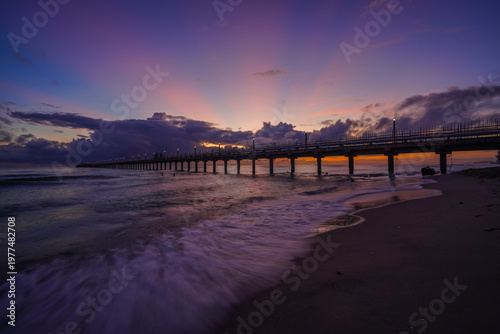 Sunset light spreads across the sky as a long pier stretches into the sea, with gentle waves reflecting purple and gold tones along the shore.