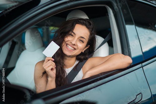 Happy cheerful young woman showing a driving license while sitting in the modern car. Driving test, driver courses, exam concept