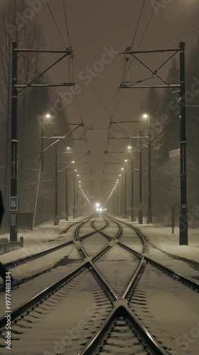 A sepia-toned winter night scene of two people walking along a snow-covered railway track, with overhead power lines and streetlights illuminating the misty atmosphere