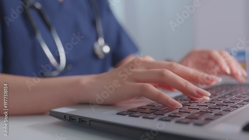 A female doctor is typing on a computer keyboard at a white wooden desk in her hospital office. The image shows a close-up of her hands, which are recording diagnoses and patient symptoms.