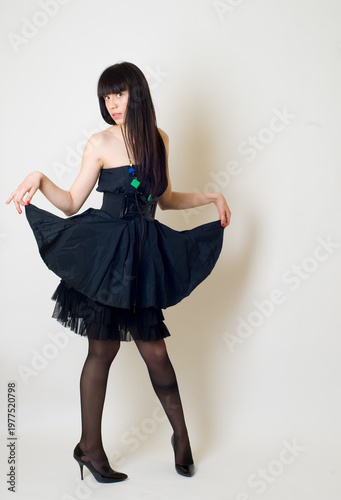 Studio portrait of a young woman wearing an elegant black dress, posing against a clean white background. The model is captured in a full-length or three-quarter pose, exuding confidence