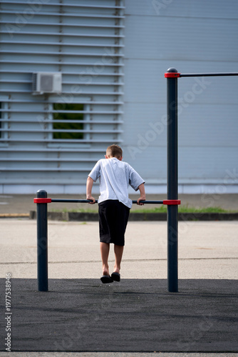 Little schoolboy, young athlete is pulling himself up on a sport horizontal bar on outdoor playground on sunny day. Active childhood, acrobat, workout on backyard, summer camp concept