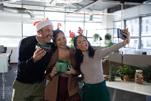 Diverse coworkers wearing Santa hat and antlers, taking selfie with smartphone and gifts in office