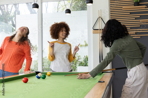 Three adult female friends playing pool on green table with cues and balls in recreation room