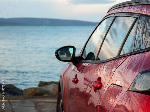 Red car parked in car park with stunning view on nature scenery with ocean and mountains and cloudy sky. Silverstrand area, Galway city, Ireland. Rent motor for a tourist trip concept.