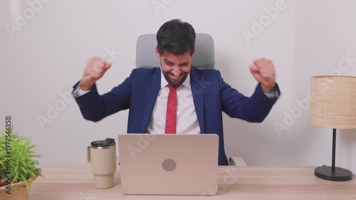 Excited young businessman using a laptop computer, celebrating success, a good deal, or a win by raising his fists in his modern office. Corporate employee feeling euphoric after an achievement