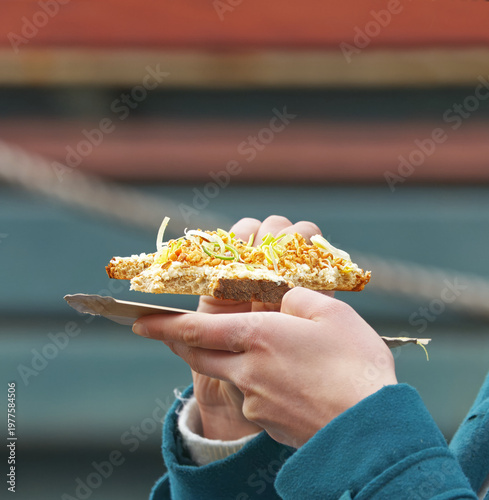 Close-up of hands holding a delicious artisan open sandwich with crispy onions on a paper tray at the popular Naplavka farmers market in Prague. Great for street food and local lifestyle concepts.