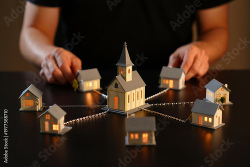 Person arranging miniature houses and church with string lights on a dark table indoors