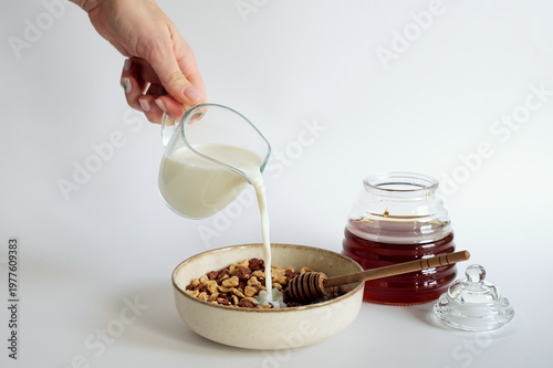 Female hand pouring fresh milk into a bowl of dry cereal flakes and honey.