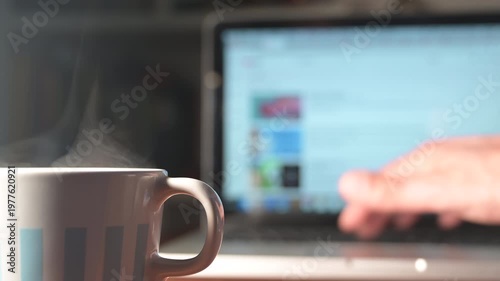 Close-up video of a steaming coffee cup in the foreground, with a blurred laptop in the background where a hand is typing. Cozy and productive atmosphere representing remote work, daily routine