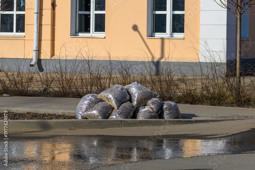 Pile of grey plastic garbage bags left on a city sidewalk next to a puddle with reflection.Spring city cleanup results: sacks filled with waste and dry leaves waiting for disposal.