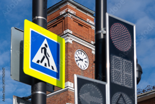 Street view of a classic tower clock against a red brick facade with a blurred pedestrian crossing sign in the foreground.