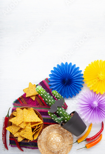 Cinco de Mayo festive composition with tortilla chips, chili peppers, cactus with sombrero and mustache and traditional Mexican textile on white wooden background