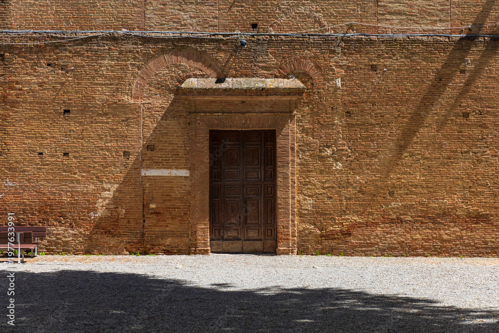 Fototapeta premium Historic Wooden Door in Sunlit Brick Wall, Siena, Italy