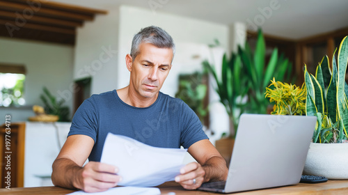 Man concentrating on paperwork and laptop, handling finances and working from his home office setting