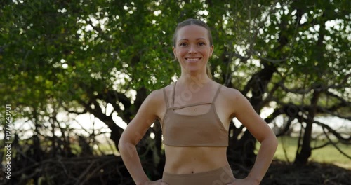 Portrait of positive sportive woman in activewear holds hands on hips as stands in green park. Pretty sportswoman radiates confidence at outdoor training