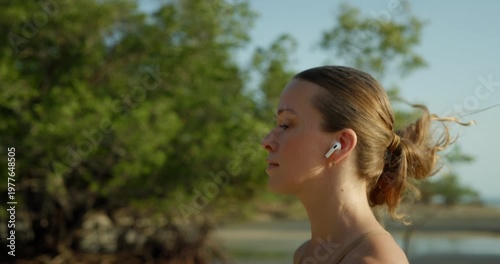 Portrait of tranquil woman with earphones runs on ocean beach with growing palm trees. Young fair-haired lady athlete exercises jogging at sunny morning seaside