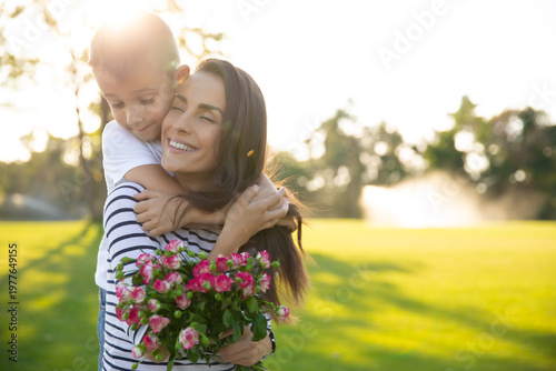 Smiling mother with a bouquet of flowers hugged by her little son outdoors, warm family moment, love, gratitude and Mother’s Day concept.
