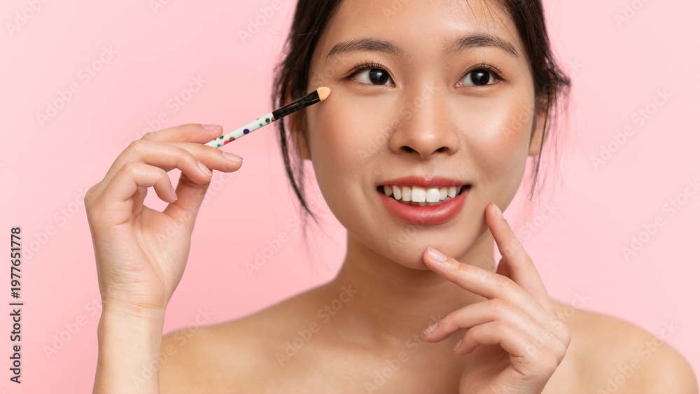Fototapeta premium A woman holds a makeup brush near her face while smiling. She has a playful expression and is focusing on her makeup application. The background is a soft pink color.