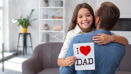 Happy daughter hugging her father and holding a handmade Father’s Day card at home. Family love, bonding, care, appreciation and holiday lifestyle concept.