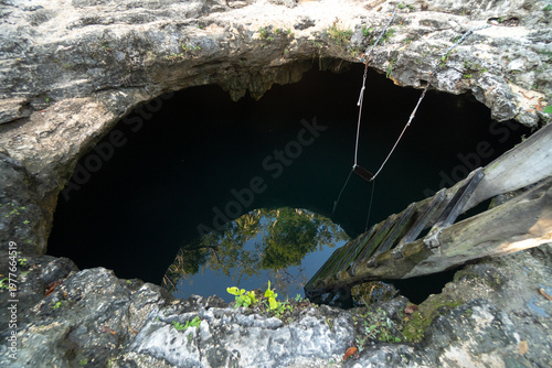 A natural cenote with a wooden ladder and swing in Yucatán Peninsula Mexico. Daytime view with rocky surroundings and water reflection.