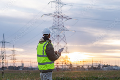 A young male engineer checks high-voltage power lines with a tablet outdoors during sunset, focusing on safety and energy management.