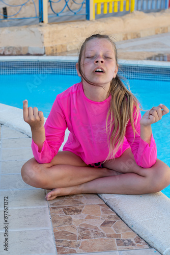 A girl in a bright bathing suit sits by the pool and meditates. A teenager sits cross-legged in a yoga pose with his arms raised, selective focus.