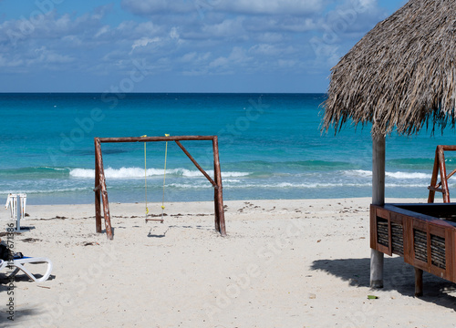 A photograph of a beach landscape: white sand, turquoise ocean with small waves, blue sky with clouds. In the foreground is a wooden swing mounted on a frame, and part of a straw beach canopy (palapa)