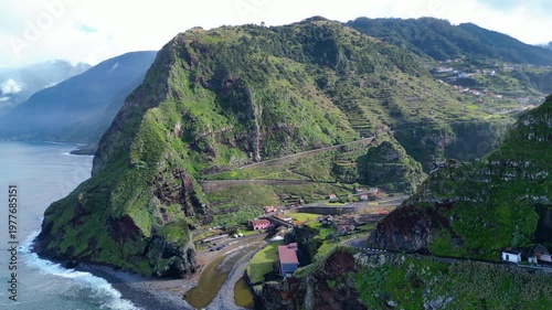 An aerial view captures the dramatic green cliffs and winding coastal road of Madeira, Portugal, near Seixal, where mountains meet the Atlantic.