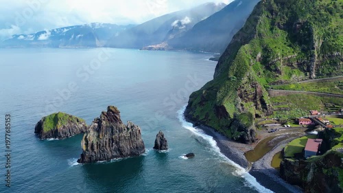 Dramatic aerial view of Madeira's rugged north coast, featuring lush cliffs, a winding road, and the iconic sea stacks of Ribeira da Janela.