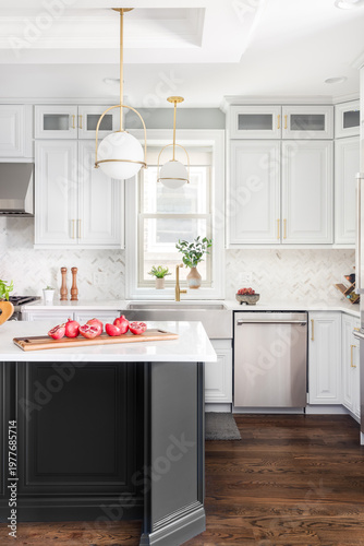 A kitchen detail with gold pendant lights, stainless steel apron sink, white cabinets and large dark grey island, and herringbone marble backsplash.