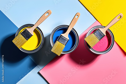 Top view of three open paint cans in blue, yellow and pink with two wooden paintbrushes on geometric color-blocked background