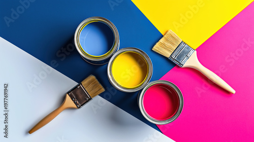 Top view of three open paint cans in blue, yellow and pink with two wooden paintbrushes on geometric color-blocked background