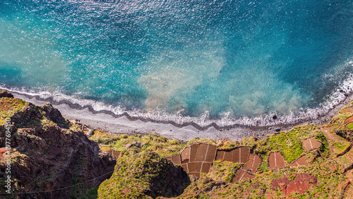 Aerial view of rocky coastline with turquoise ocean and terraced fields. Dramatic cliffs meet clear water, showing natural beauty and cultivated land