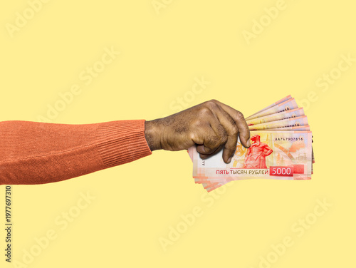 A hand in an orange sweater holds a fan of 5000-ruble bills against a yellow background. A clean, conceptual image representing Russian currency, wealth, spending, and financial success.