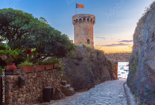 Schilderij op canvas Historic stone watchtower overlooking the calm bay of Tossa de Mar, Costa Brava, Spain, with anchored boats and warm sunset light over the Mediterranean coast
