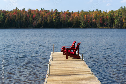 Two red Adirondack chairs with matching cushions sit at the end of a floating dock on a blue Ontario lake, facing a dense wall of peak autumn foliage across the water.