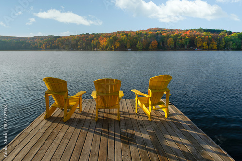 Three bright yellow Muskoka chairs are arranged side by side on a wide wooden dock, all facing a large Ontario lake at the golden hour of a fall morning. A rolling hillside of vibrant red, orange,