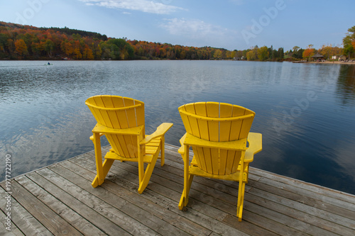 Two bright yellow Muskoka chairs face a wide Ontario lake bay from a weathered wooden dock, with rolling hills of vibrant peak autumn foliage curving around both shores on a partly cloudy fall day.