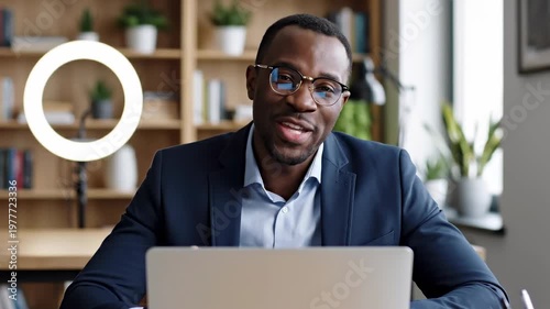 Smiling black businessman on laptop webcam call in modern home office, talking to colleagues during remote work video meeting with ring light and bookshelf background indoors at desk