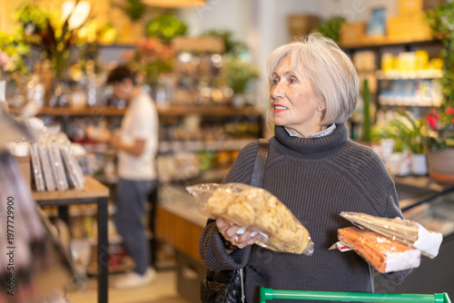 Store visitor, senior woman in sales area choosing pasta. Client make choice of assortment, select, buy noodle product. Customer read label, examine customer information, view package content
