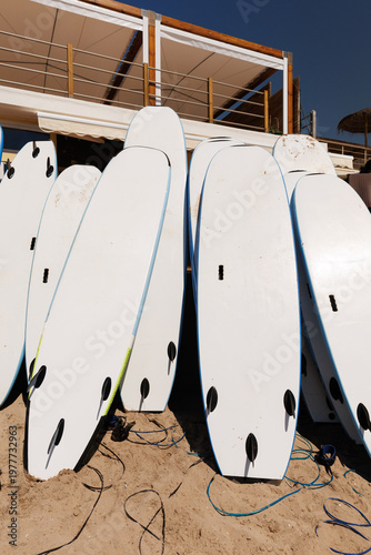 Photography A row of white surfboards with black fins leaning against a structure on a sandy beach, prepared for surfing lessons or rentals