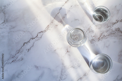 Glass of water under sunlight creating abstract shiny reflection on marble floor. Minimal aesthetic top view summer lifestyle.