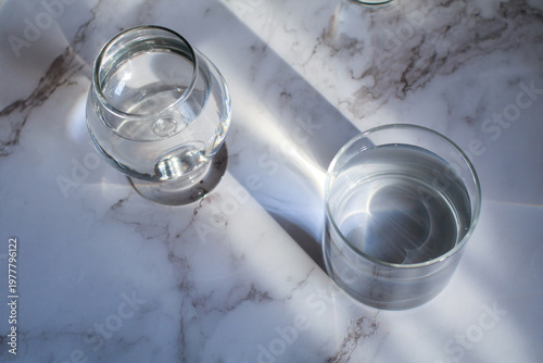 Glass of water under sunlight creating abstract shiny reflection on marble floor. Minimal aesthetic top view summer lifestyle.