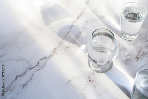 Glass of water under sunlight creating abstract shiny reflection on marble floor. Minimal aesthetic top view summer lifestyle.