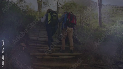 Hiking couple climbing log steps at trail start, planting pole and carrying backpacks to ridge