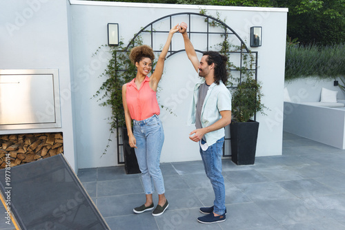 Diverse couple dancing on tiled patio at home near trellis and built-in grill, wearing casual shoes