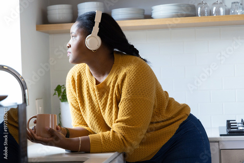 African American woman leaning at sink wearing mustard knit and white headset holding peach mug