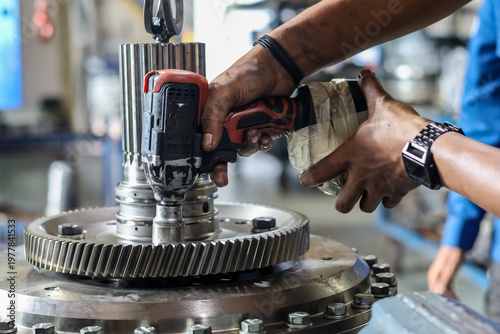 Photos Technician using electric cordless impact wrench to tighten bolts on heavy metal gear in industrial workshop