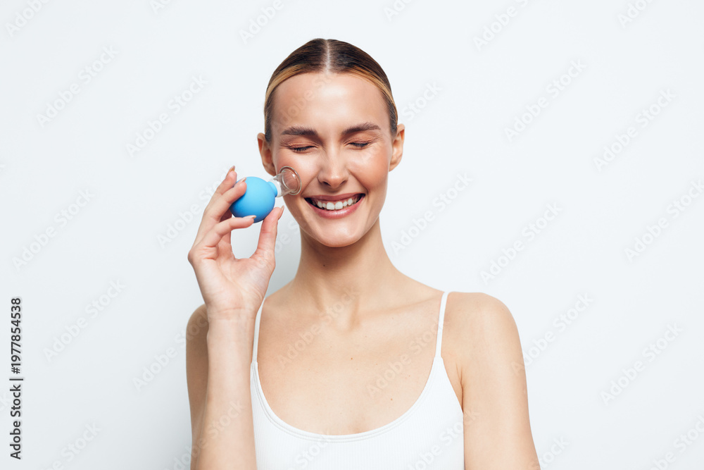 Naklejka premium A smiling young woman with closed eyes holds a blue facial cleansing sponge near her cheek, demonstrating skincare routine and beauty care on a white background.